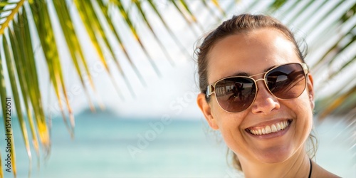 Smiling woman wearing sunglasses on a tropical beach. Palm tree leaves in the foreground, ocean in the background. Happiness, summer vacation vibes