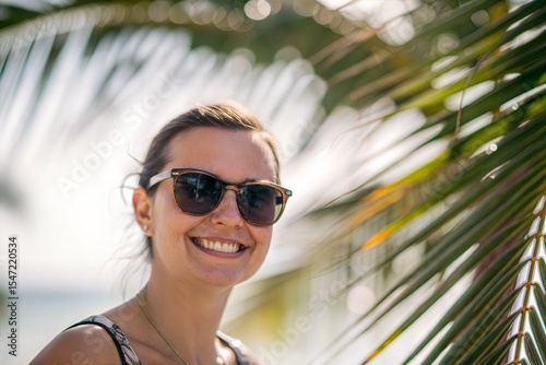 A smiling woman wearing sunglasses in the sun, with palm leaves framing her face. A vibrant moment of happiness and warmth.