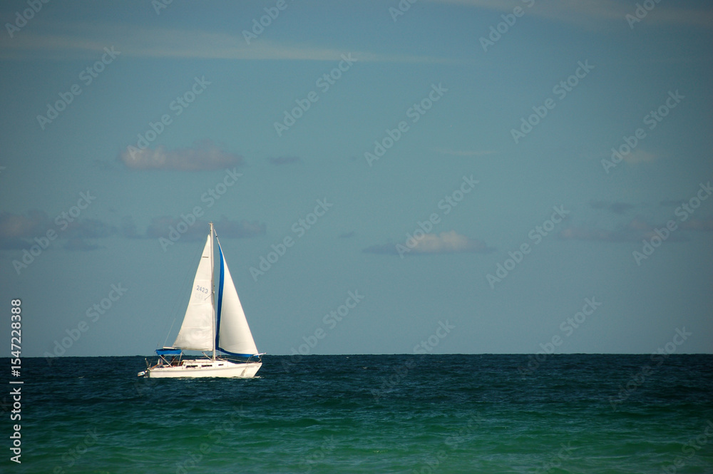Fototapeta A white sailboat with blue accents sails peacefully on the turquoise ocean under a partly cloudy sky, creating a serene seascape