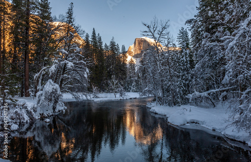 The peak of half dome is baking in the full light of the setting sun. It is reflected in the merced river, on a bright snowy winter afternoon during golden hour.