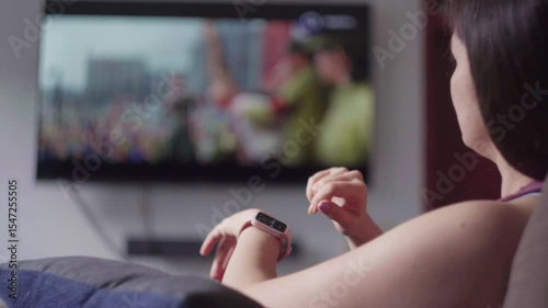 Woman relaxing on sofa, browsing smartwatch screen during tv program, representing contemporary connected digital living