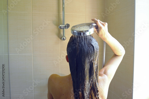 Self-care routine at home: a woman, photographed from behind, stands in a bathroom, head thrown back, rinsing her reddish-blond hair under a stream of water from a shower head she holds in her hand.