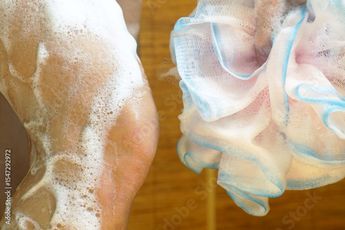Close-up of the shower process: a woman's bent leg covered in shower gel foam and a voluminous synthetic sponge. Daily hygiene: keeping the skin clean.