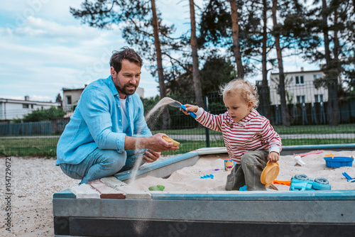 Father and little toddler son playing in the sandbox on playground.