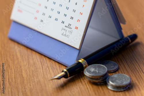 Fotografi A desk calendar with pay off debt! marked on a date beside a pen and a few stacks of coins
