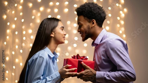 Young diverse couple exchanges gift and kisses against romantic bokeh lights footage. Representing love, anniversary, Valentine's Day or holiday celebration.