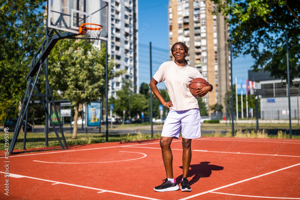 Fototapeta premium Young woman practicing basketball alone at sports court in the morning. Happy girl is ready for playing basketball on sunny day.