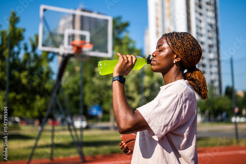 Young black woman practicing basketball alone at sports court in the morning. She is drinking energy drink.