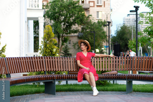 Woman in red dress and hat poses thoughtfully on park bench.