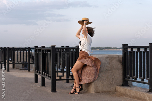 Wind lifts skirt as woman holds straw hat by river