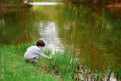 young boy exploring nature by pond, crouching amidst lush green grass, reflecting childhood curiosity and outdoor learning. serene atmosphere enhances peaceful exploration vibe