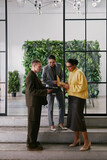 Vertical shot of two young adult women and man standing together in modern office discussing business documents, collaborating on project near glass wall with greenery
