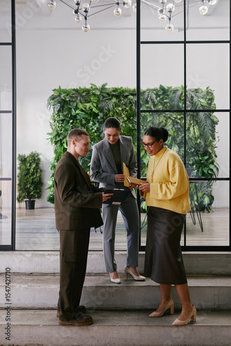 Vertical shot of two young adult women and man standing together in modern office discussing business documents, collaborating on project near glass wall with greenery