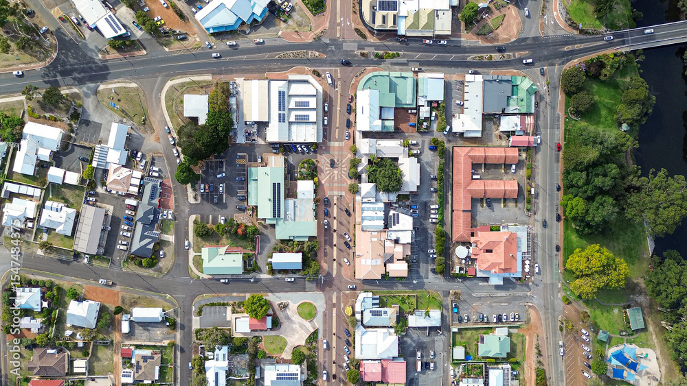 Φωτογραφία Top down Arial view of a small rural town centre with cars on the road, roof tops of homes and businesses, a river with park and trees