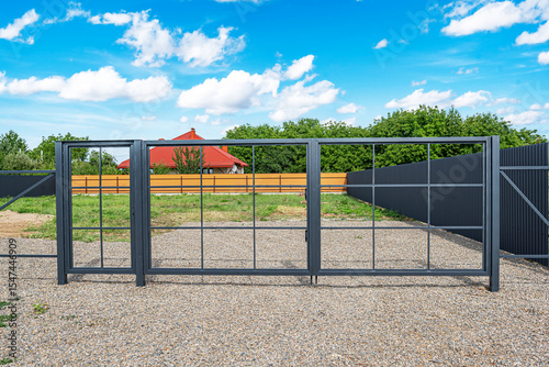 A modern metal gate with a grid design stands closed on a gravel driveway, leading to a fenced residential property under a bright blue sky.