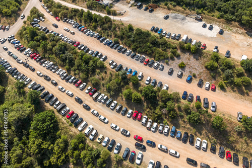Fototapeta Naklejka Na Ścianę i Meble -  Top-down view of a large dirt parking area filled with neatly arranged cars in a Mediterranean forest near a beach resort, showing tourism infrastructure behind the scenes