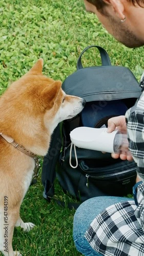 Young man in plaid shirt giving water to his dog from a special bottle during outdoor walk summer park pet care hydration concept companionship	
