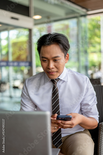 Male office worker holding phone and looking at laptop on table sitting in a cafe or coffee shop.