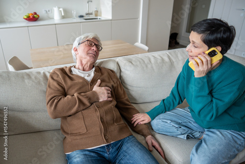 Concerned wife makes urgent call during husband heart attack. Senior male collapsing, chest pain, panic in room, aging health risk, cardiovascular failure, immediate assistance and domestic emergency