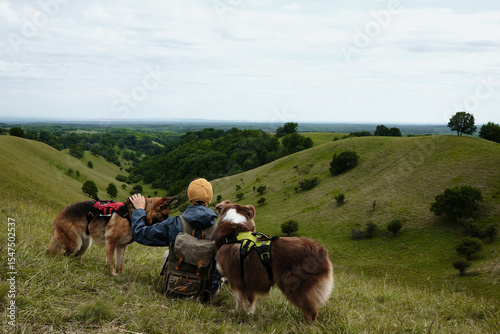 Woman with two dogs sits on the edge of a hill, watching a picturesque valley in Serbia. Rear view. Zagajicka Brda in Serbia in spring season. German and Australian Shepherd travel with owner