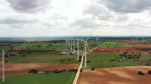 Aerial drone view of multiple wind turbines rotating over colorful farmland under cloudy sky. Scenic footage highlights clean energy production and sustainable environmental practices