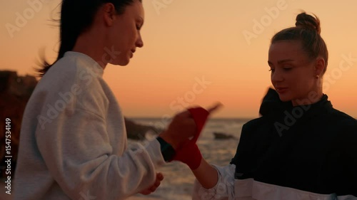 Trainer removing boxing tapes from woman's hands after workout near ocean during sunset, showing care, discipline, and post-training routine