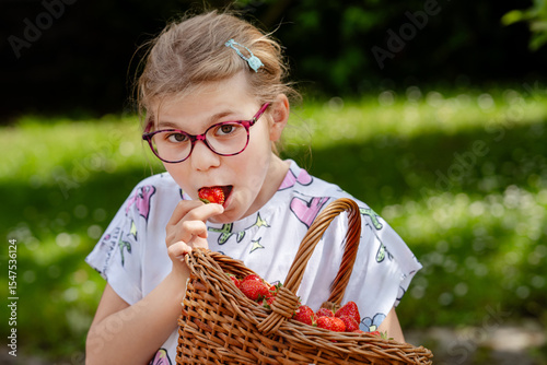 Happy little school girl holding a big basket full of ripe, sweet strawberries. Real life, real people moment of childhood joy and summer fun outdoors in nature.