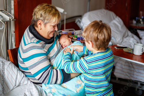 Happy grandmother holding newborn baby in hospital with proud big brother nearby. Real life, real people moment of family love, siblings, and multigenerational bonding.