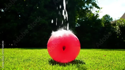 Pink water balloon bursting and splashing on green grass in sunlight  