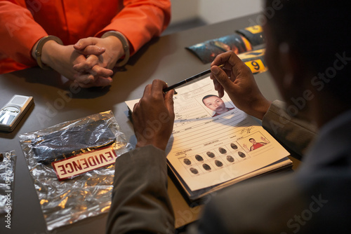 Middle aged Black man interrogating middle aged Caucasian man in orange prison uniform at table with evidence bags, crime scene photos, and police report featuring fingerprints and mugshot