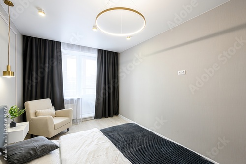 modern bedroom features a bed, armchair, dark curtains, and a striking circular ceiling light. Neutral color palette with accents of gold in lighting