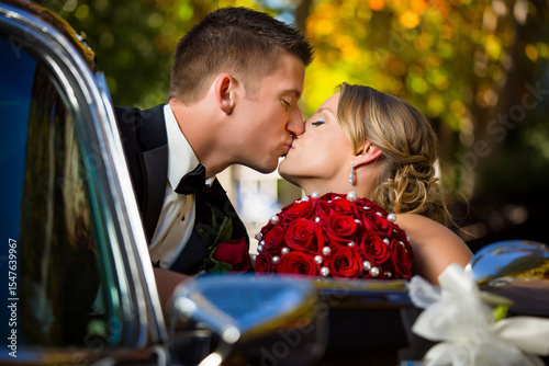 Closeup of a wedding couple kissing beside a classic car, holding a red rose bouquet with pearls. Romantic outdoor portrait with bokeh autumn trees in the background.