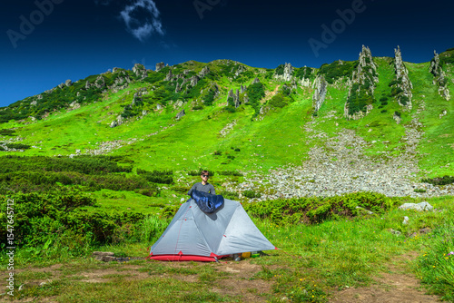 Camper drying sleeping bag on tent in green mountain campsite. Summer outdoor adventure, trekking and wilderness morning routine