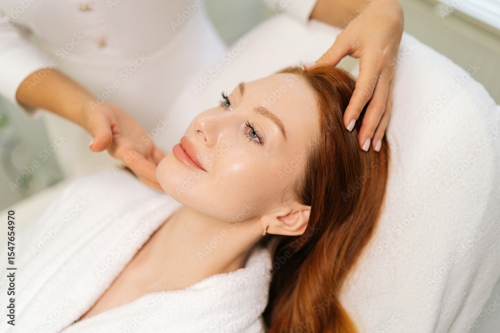 Close-up of beautician performing relaxing facial massage for young woman with long red hair, lying on massage table in white bathrobe, enjoying skincare treatment in modern beauty salon. Close-up of beautician performing relaxing facial massage for young woman with long red hair, lying on massage table in white bathrobe, enjoying skincare treatment in modern beauty salon.