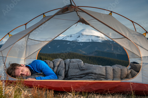 Man sleeping under down quilt inside tent during early mountain sunrise. Outdoor camping and morning wilderness scene