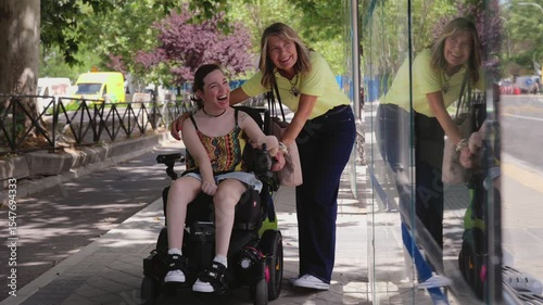 Disabled woman in electric wheelchair sharing hearty laughter with friend at urban bus stop, glass reflection capturing genuine moment of joy and connection