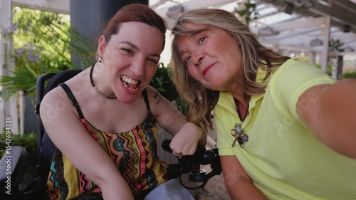 Disabled woman in wheelchair capturing joyful selfie with her mother, sharing warm smile and connection during outdoor moment