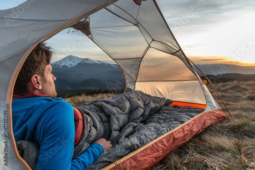 Man lying in tent with sleeping bag, watching mountain sunset view, camping in the wilderness, outdoor adventure in nature