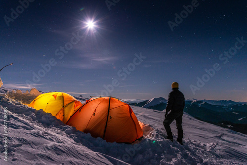 Man standing by winter tents on snowy mountain, under starry night sky and bright moonlight, alpine camping scene