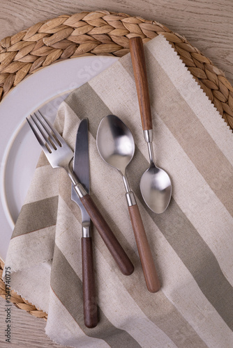 A modern cutlery set with wooden handles is elegantly arranged on a striped linen napkin atop a wicker placemat. The set includes a fork, knife, and two spoons, highlighting contemporary dining style.