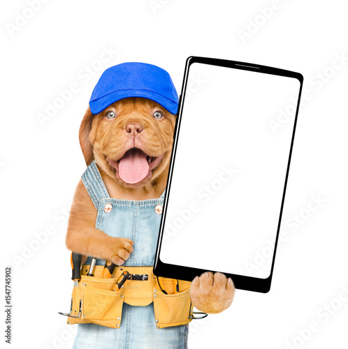 Happy Mastiff puppy wearing blue cap and overalls with tool belt showing big smartphone with white blank screen in it paw. Isolated on white background