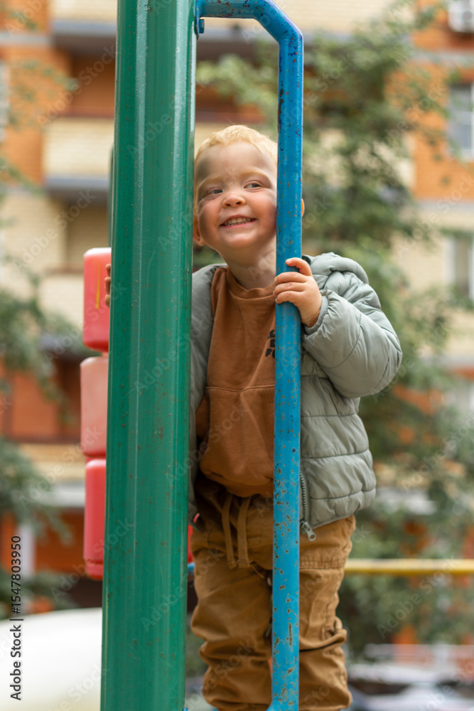 Obraz premium Vertical portrait of a small red-haired boy who looks over the fence of a playground in the city. Outdoor games