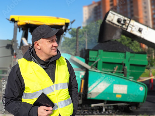 Man foreman of road construction company. Guy near asphalt paving equipment. Contractor of road construction bureau. Man foreman supervises road construction. Worker in yellow vest build highway
