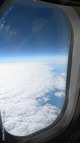 View from inside an airplane cabin window looking out at a vast white cloudscape below a vibrant blue sky, dark interior frame highlights bright aerial perspective as aircraft flies high above earth,