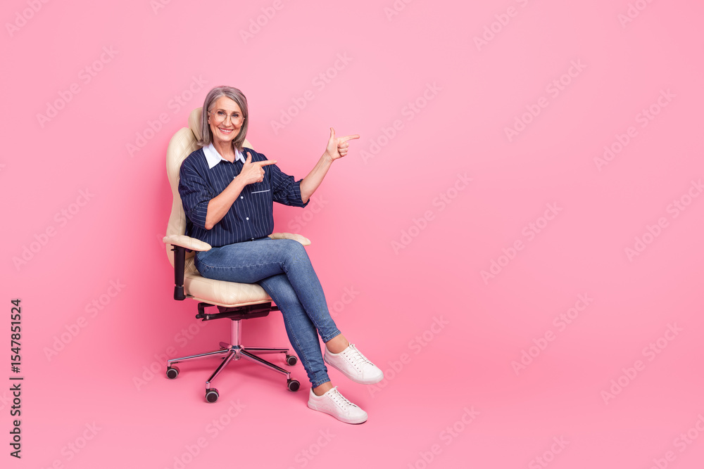 Mature businesswoman in stylish casual outfit sitting on chair against pink backdrop, pointing confidently to the side Mature businesswoman in stylish casual outfit sitting on chair against pink backdrop, pointing confidently to the side