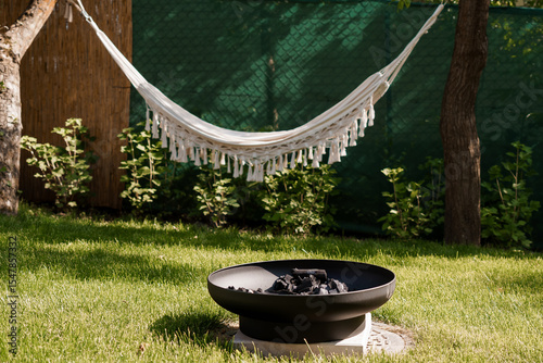 Cozy Backyard Retreat with White Hammock and Fire Pit Scene: Relaxing Outdoor Living, Green Garden, Summer Comfort, and Dark Metal Fire Bowl.