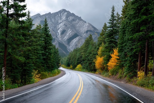 A winding road leads to a tall mountain surrounded by green trees under a cloudy sky