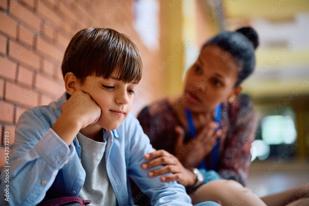 Obraz premium Sad schoolboy with supportive teacher in school hallway.