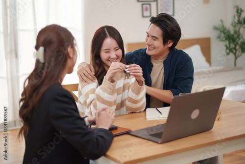 Happy young Asian couple receiving new house keys from female agent at home, smiling and celebrating success, concept of home ownership and real estate investment