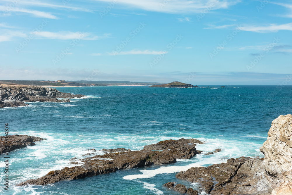 Fototapeta premium View of the rocky coastline near Ilha do Pessegueiro, Portugal. Blue Atlantic waters crash against the rugged shore under a bright sky. Perfect seascape for travel and nature themes.